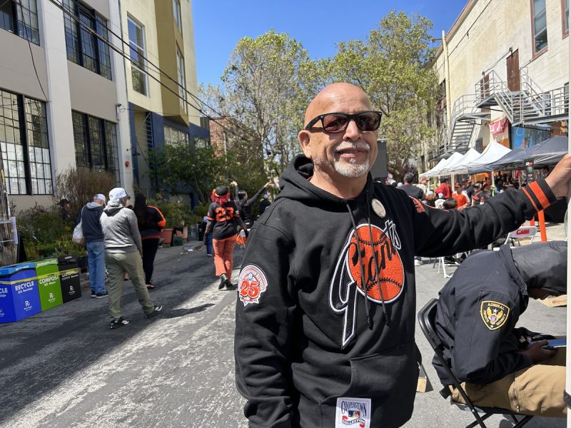 A man in sunglasses and a Giants sweatshirt poses while dozens of fans watch a game behind him.
