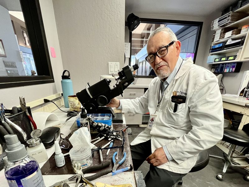 A man in a white lab coat sitting at a table with glasses.