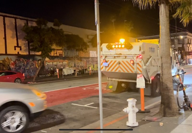 The hood of a car is poking out as it drives into a parking spot behind a street sweeper.