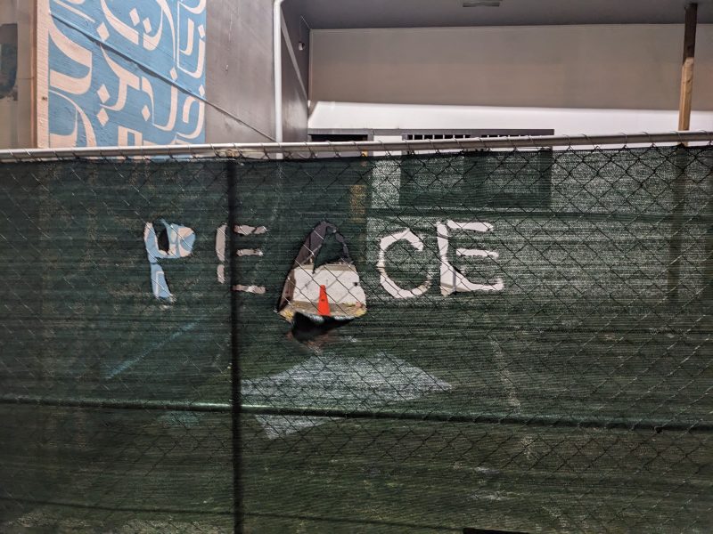 Graffiti on a fence screen with the word PECE. the A has been torn out.