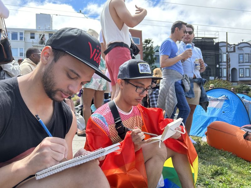 Two queer people, one wrapped in a Pride rainbow flag, sketching.