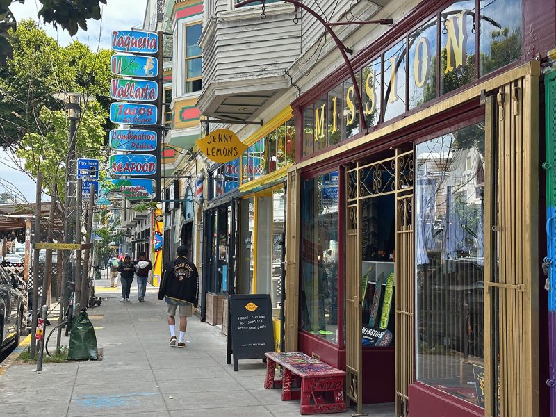 An urban street with storefronts and a few pedestrians walking by