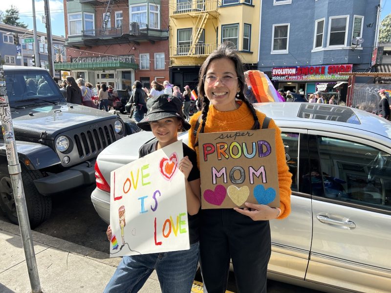 A mother in a bright orange sweater carries a sign that says 'Super Proud Mom' next to her daughter, in a bucket hat, that carries a sign that says, 'Love is Love.'