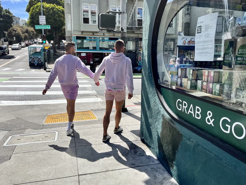 Two men dressed in pink shirts and pink shorts, from behind, hodling hands as they round the corner of a store.