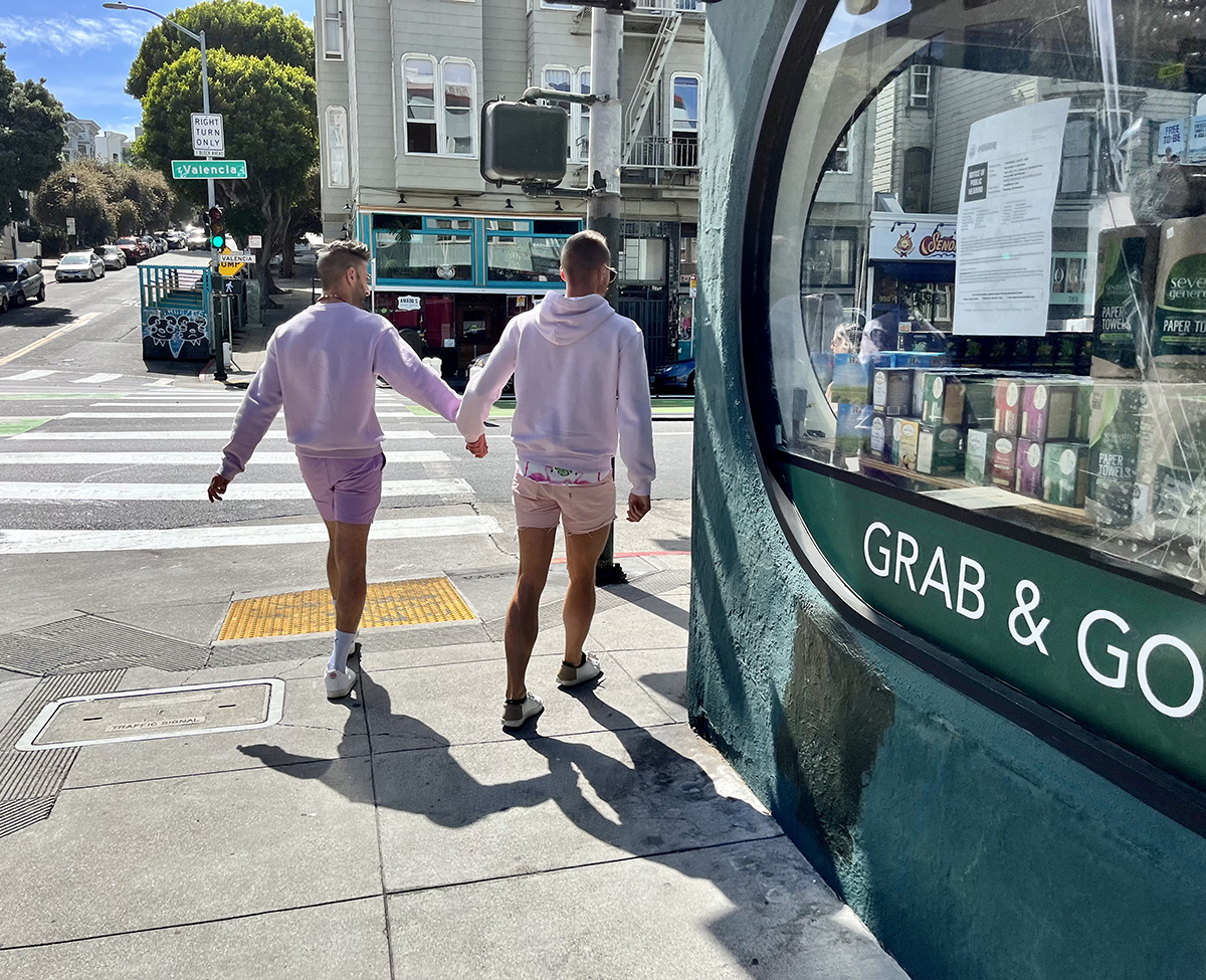 Two men dressed in pink shirts and pink shorts, from behind, hodling hands as they round the corner of a store.