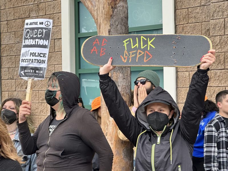 masked skateboarder holding up a skateboard reading "Fuck SFPD" in a crowd of protesters