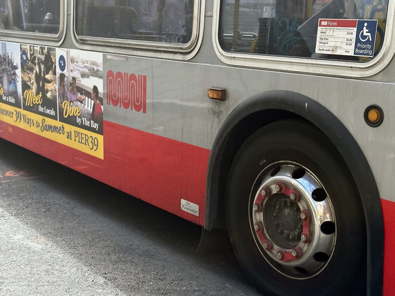 The side of a red and grey MUNI bus with one wheel and he bottom of some windows showing. Not the 22-Fillmore