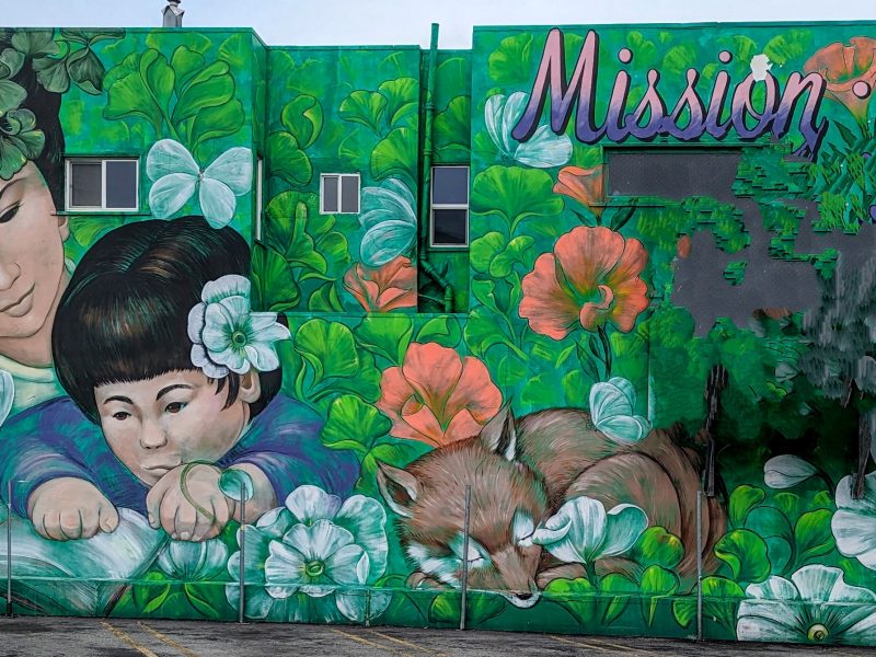 A mural of a mother looking over the shoulder of a child reading with a dog nearby, in green and some red. Lots of plants.