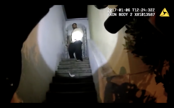 Gun points up staircase, toward a man in a spotlight standing in a doorway.