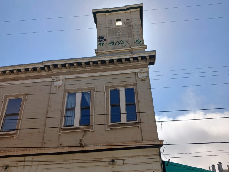 A tall building with a rectangular turret at the top with graffiti. The sky and a cloud to the right.