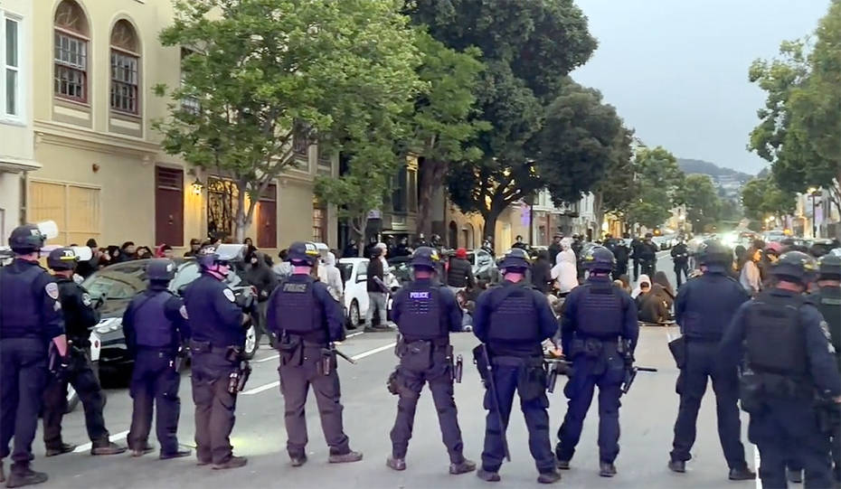 A line of police officers and between them and others a line of young skateboarders after the 2023 Dolores Park hill bomb.