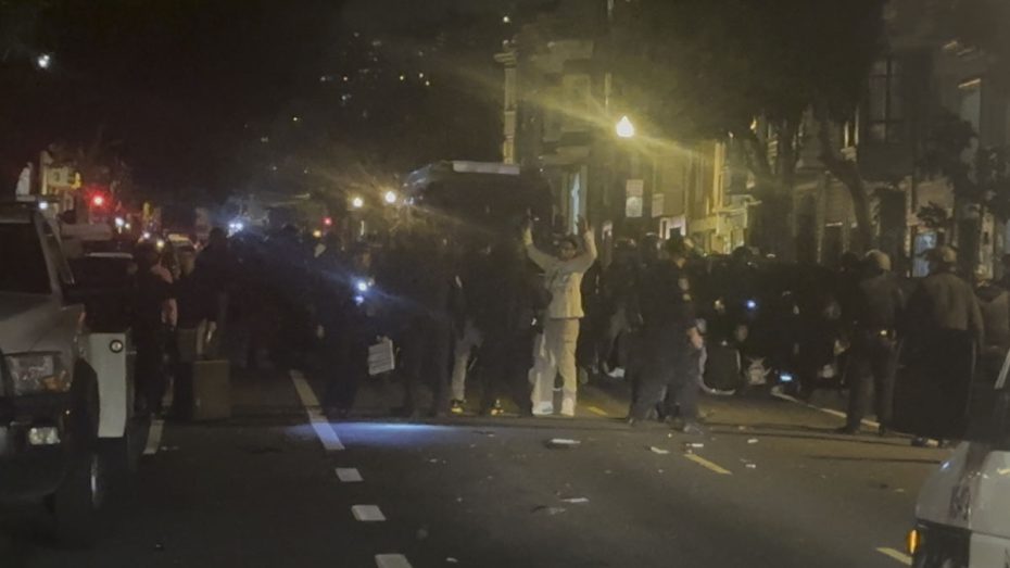 A night shot of teenagers with hands up, officers, vans after the 2023 Dolores Park hill bomb