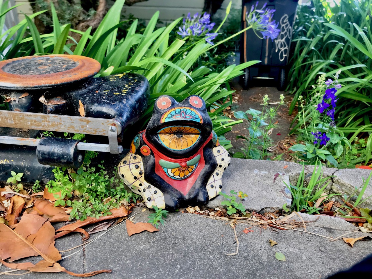 A ceramic frog, painted green, yellow and other colors sitting in a garden with green plants.
