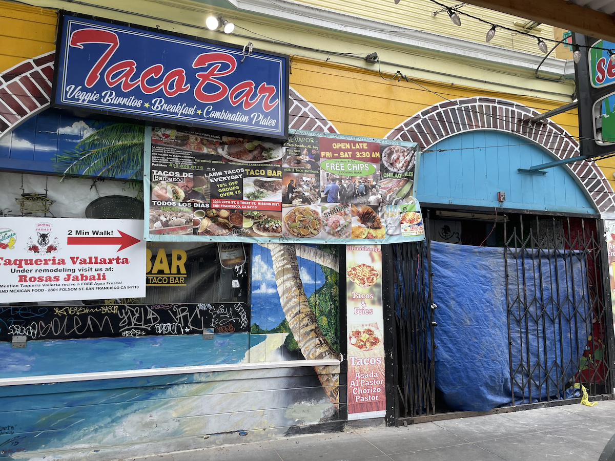 The Taqueria Vallarta storefront. A blue tarp covers the front door. A sign in the window advertises free agua fresca at Rosas Jabali on 24th and Folsom if you mention Taqueria Vallarta. Another sign advertises Vallarta's usual offerings.