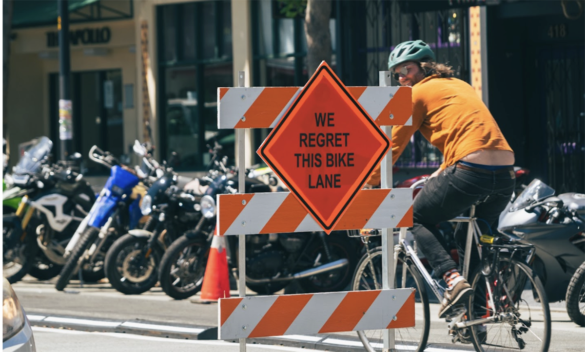 A orange sign reading "We regret this bike lane" with a cyclist riding by and parked motorcycles in the background.