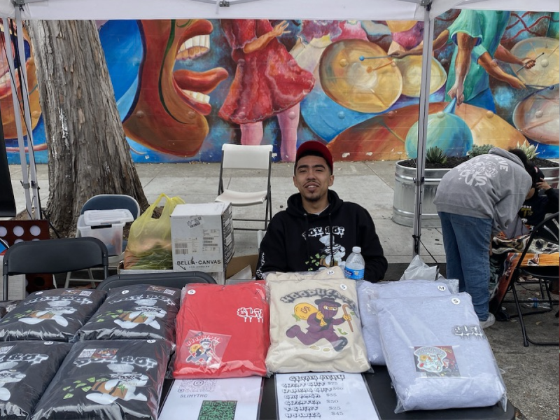 Damien Gonzalez dressed in a hoodie and a snapback mans a table full of clothes.