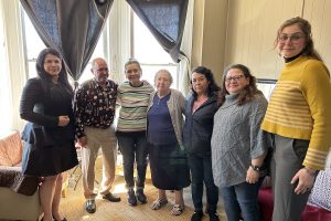 A line of seven individuals stand in front of a modest apartment with sunlit windows. A woman in a black dress and black heels is on the far most left and a young lady in a yellow striped shirt and slacks is on the far-most right. In between are a group of Latino residents, three who are seniors, who are getting evicted.