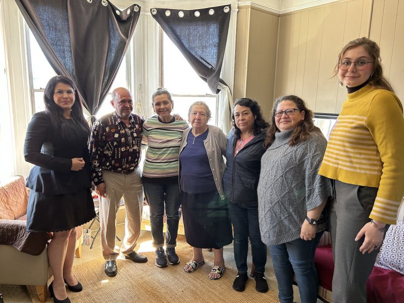A line of seven individuals stand in front of a modest apartment with sunlit windows. A woman in a black dress and black heels is on the far most left and a young lady in a yellow striped shirt and slacks is on the far-most right. In between are a group of Latino residents, three who are seniors, who are getting evicted.