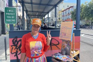 Jirsa, rainbow skirt billowing in the wind, stands in their rainbow corner, painting the buildings across the street.