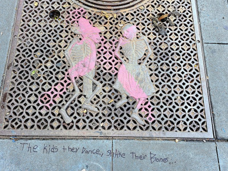 A metal grate showing two skeletons with some pink paint and the words below, the kids they dance.