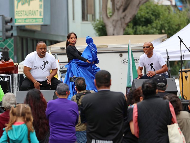 A dancer on a stage, with a blue dress on. Two musicians on either side of her.