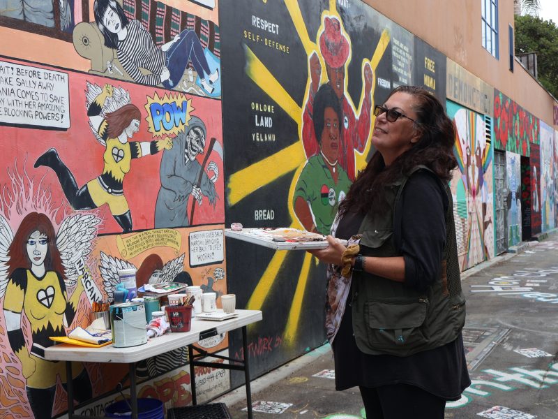 Woman holds a painting palette in her left hand, and looks up to a mural painting in front of her.
