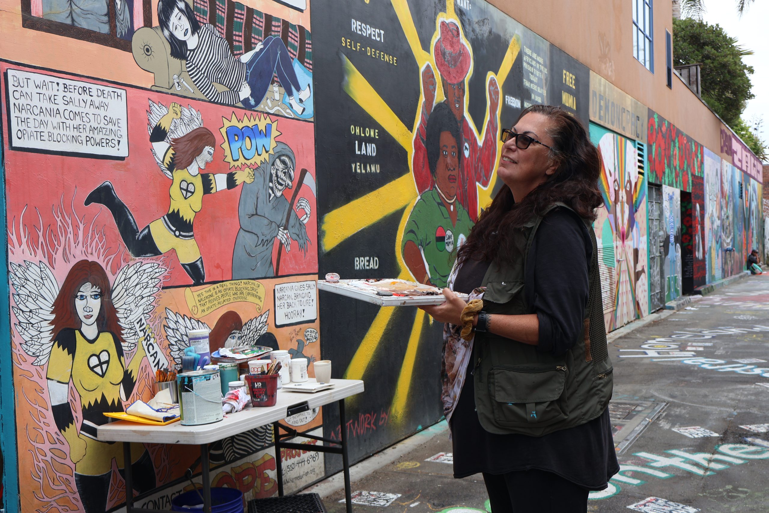 Woman holds a painting palette in her left hand, and looks up to a mural painting in front of her.