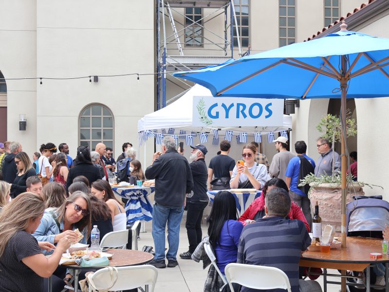 People sitting outside eating. Gyros stall in the background with people lining up to order food.