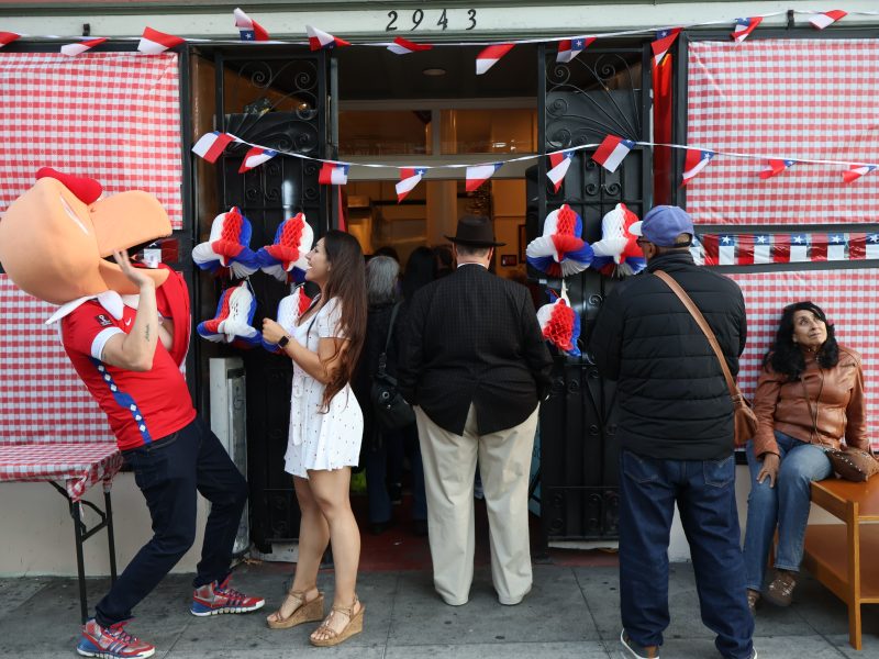 People gather outside Chile Lindo, which is decorated with Chilean flags and red and white gingham paper. A mascot, 'condorito', also stands outside.