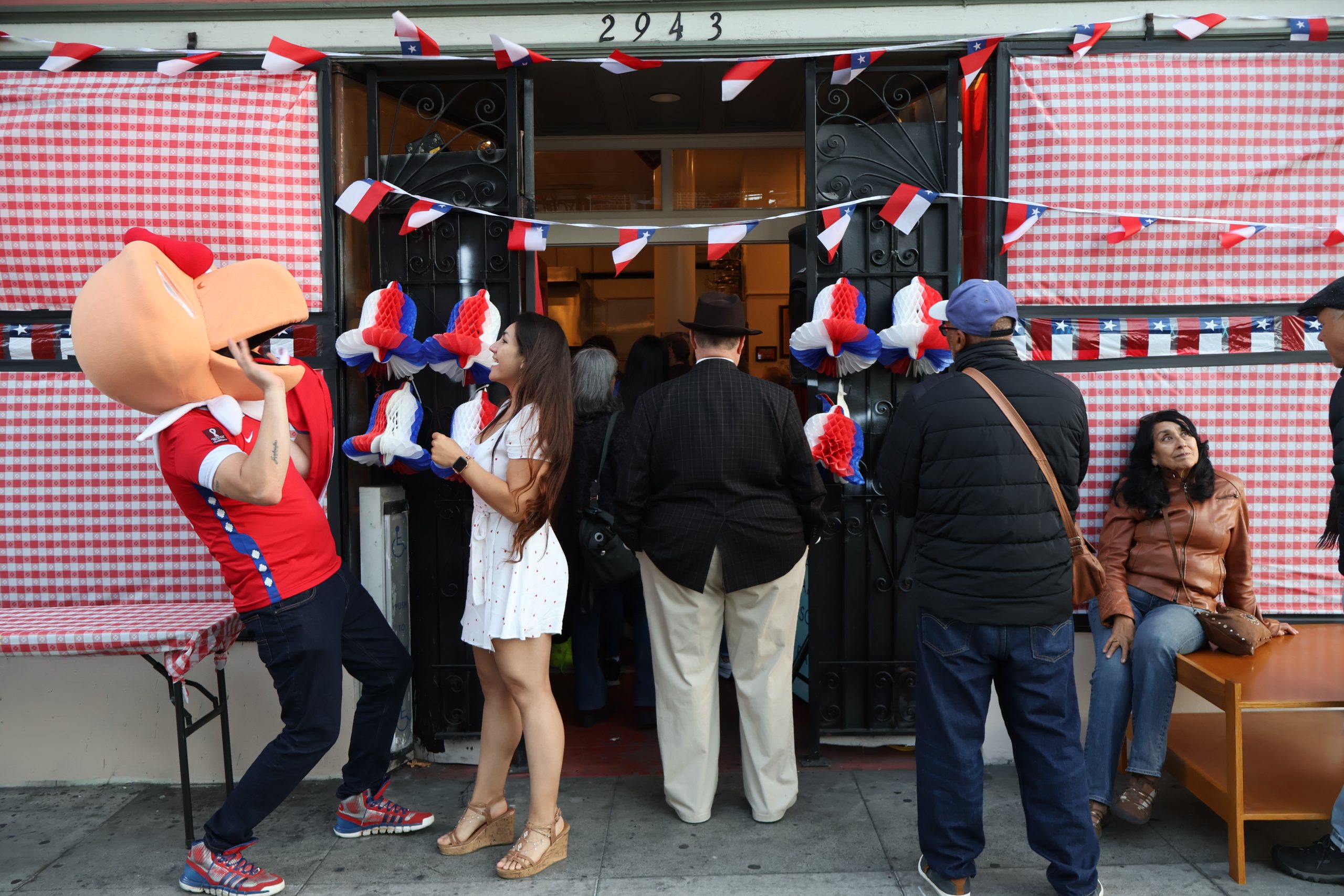 People gather outside Chile Lindo, which is decorated with Chilean flags and red and white gingham paper. A mascot, 'condorito', also stands outside.