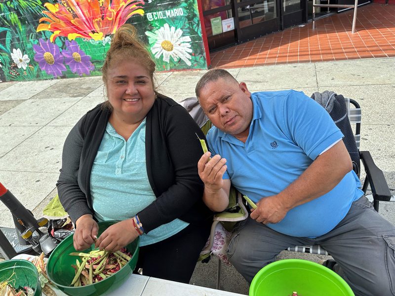 Two people, one with a black sweater and blue shirt and the other, a man in a blue shirt. They are seated and shelling beans.
