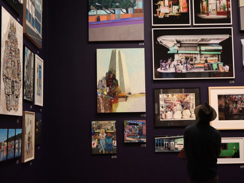 Man looks up at a crowd of paintings on a gallery wall.