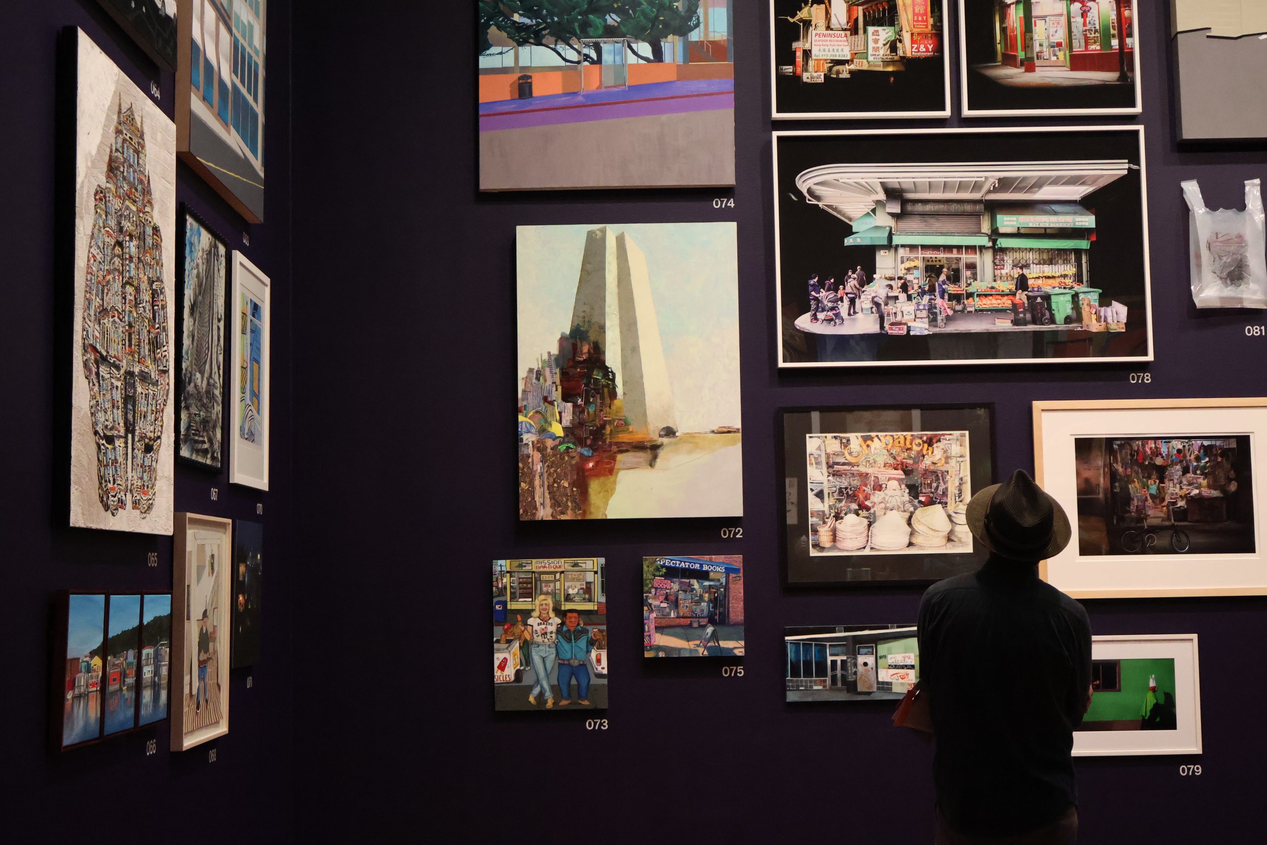 Man looks up at a crowd of paintings on a gallery wall.