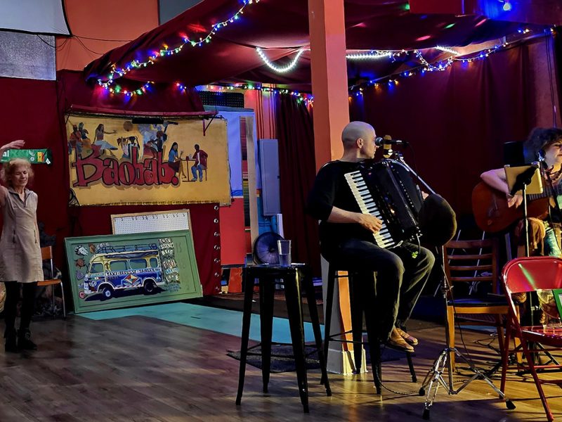 A bar scene, two people playing instruments in the foreground and a lone figure on the dance floor in the background with her arms over her head like a ballerina.