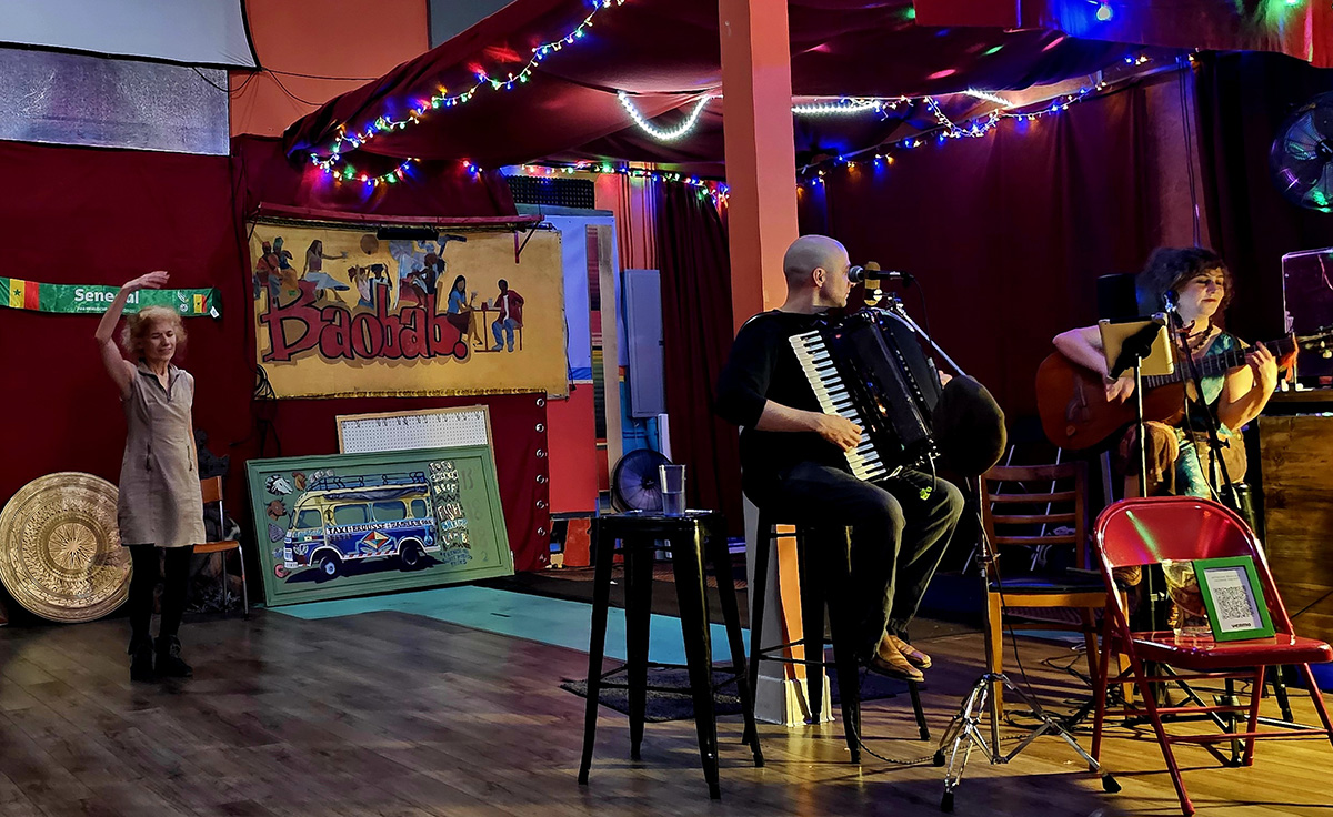 A bar scene, two people playing instruments in the foreground and a lone figure on the dance floor in the background with her arms over her head like a ballerina.