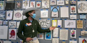A man in a cowboy hat stands in front of a wall of art.