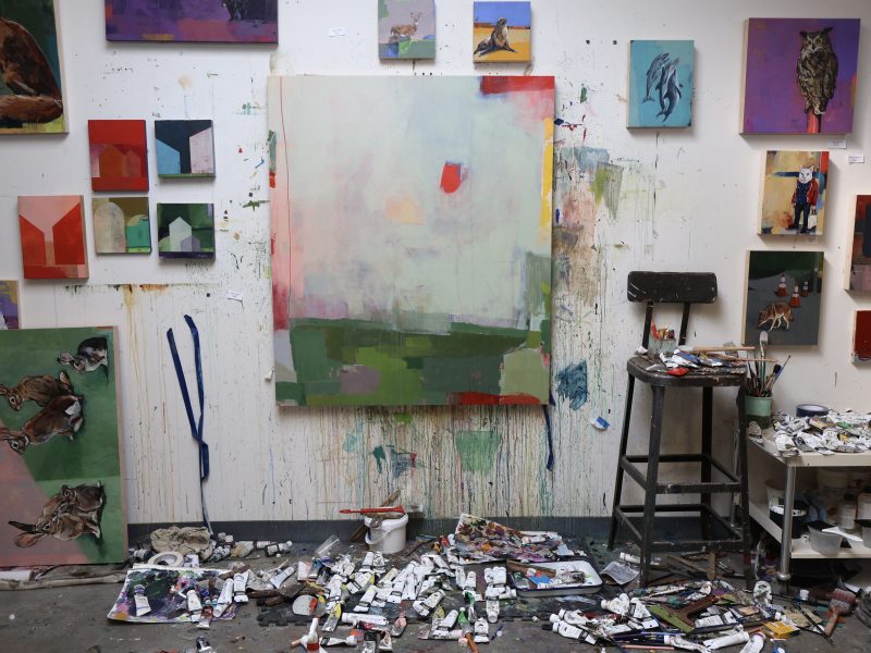 An artist's studio with a lot of paintings on the wall and paints scattered on the floor.