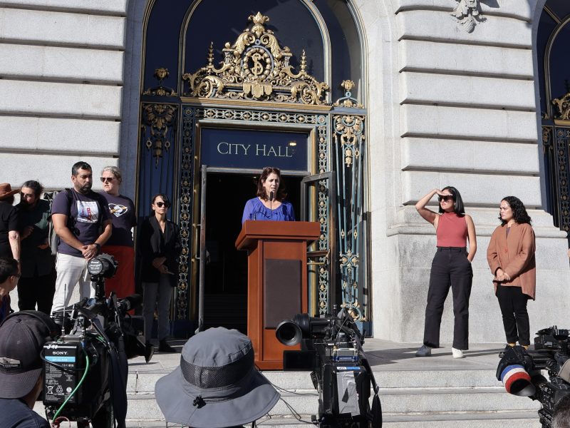 A group of people gather around a podium in front of City Hall.
