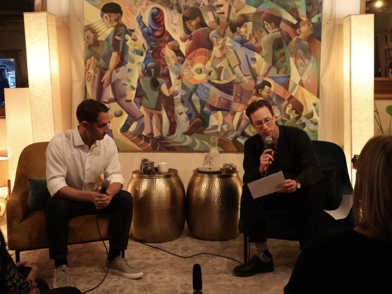 Two men sitting in chairs in front of a painting.