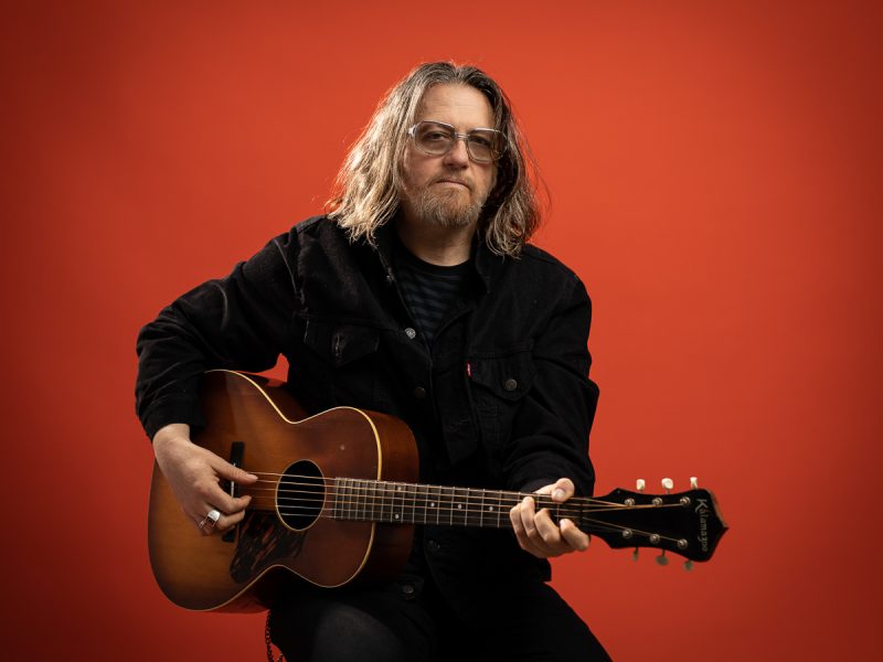 Tom Heyman holding an acoustic guitar in front of a red background.