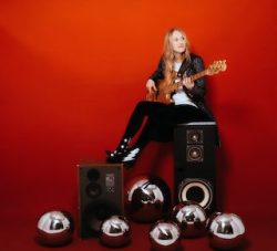 A woman is sitting on a red background with a guitar.