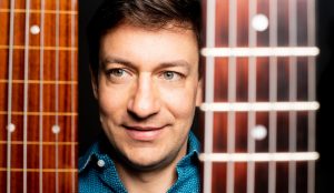 A man with a guitar in front of a black background.