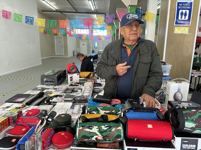 Licensed vendor Franco Gonzalez standing in front of a table full of electronics that he is selling indoors, due to the new street-vending ban