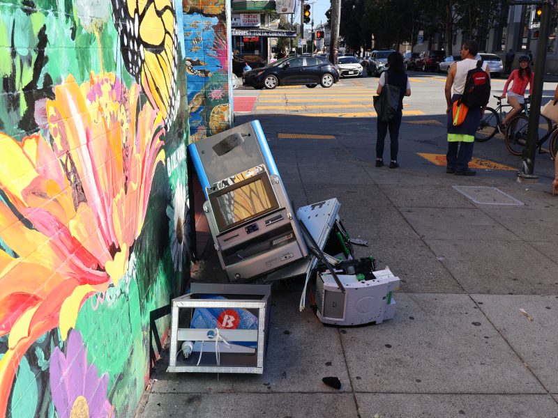 A colorful mural painted on the side of a street with parts of an ATM against it.