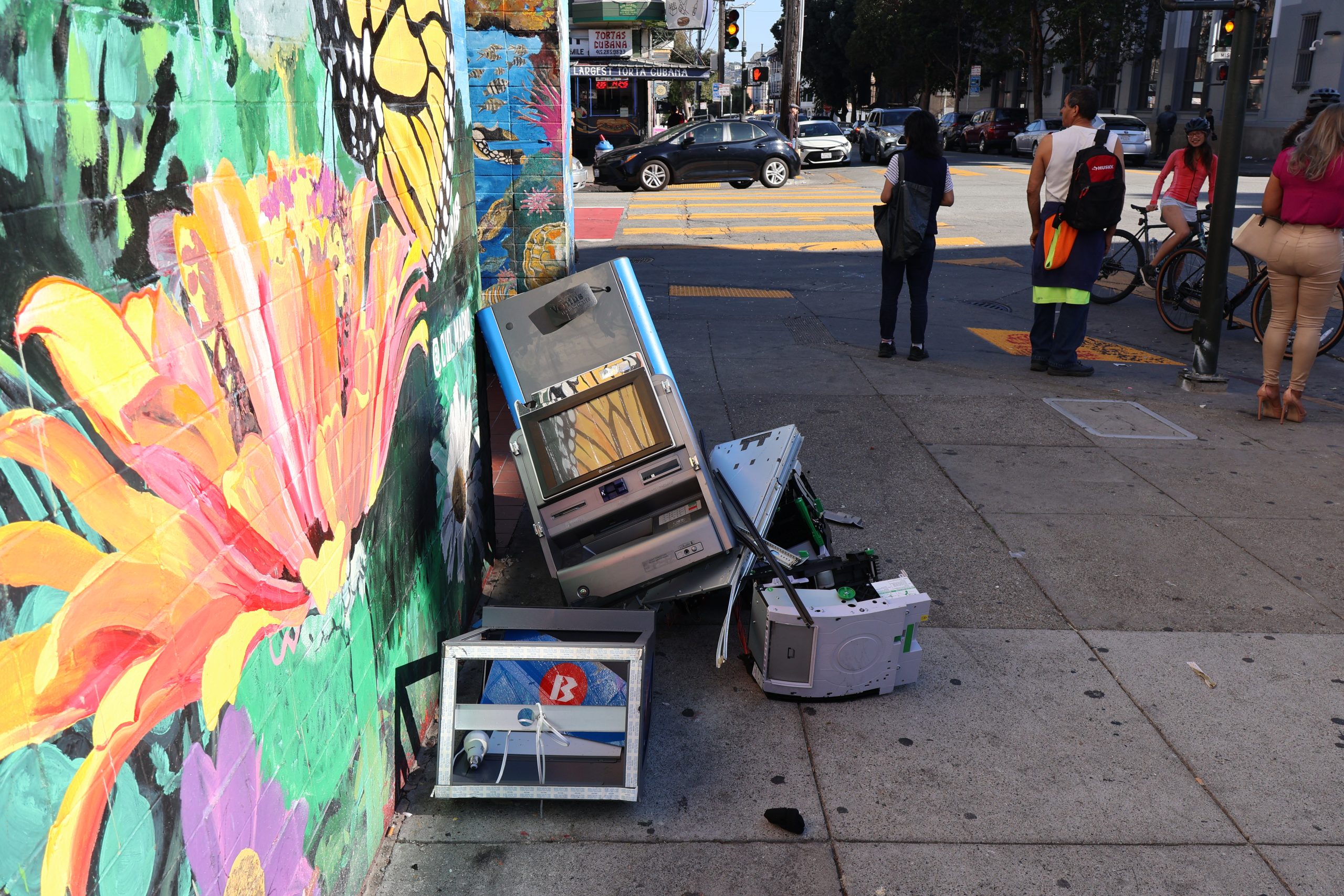 A colorful mural painted on the side of a street with parts of an ATM against it.