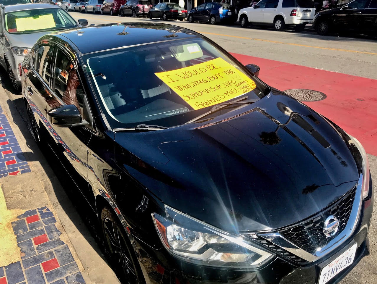 A black Nissan Sentra parked on the side of the street. A sign on its windshield says "I would be vending. But the supervisor banned me. Support street vendors."
