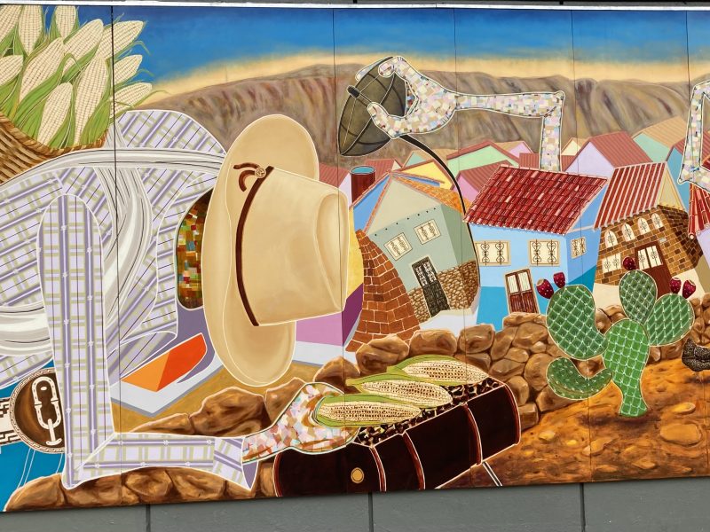 A mural on the side of a building with a man in a hat holding three cobs of corn in his hand and carrying a basket of corns on his back.