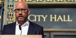 A bald man speaking at a podium in front of a city hall sign.