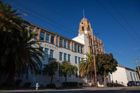 Street-level view of a historic school building with a decorative tower, palm trees, and a clear blue sky.