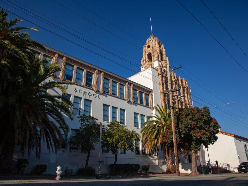 Street-level view of a historic school building with a decorative tower, palm trees, and a clear blue sky.
